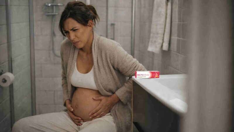 Trudnica sedi u kupatilu i drži stomak, uz vidljiv gel za hemoroide kao simbol olakšanja simptoma
