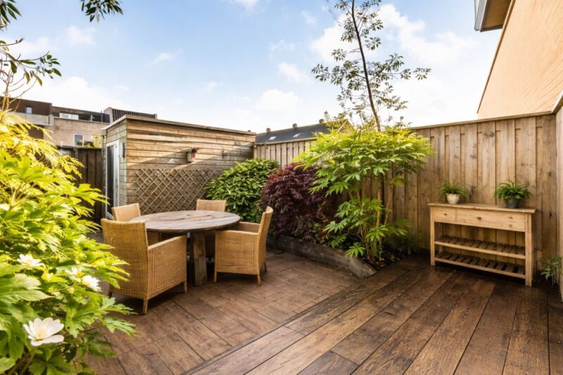 Malo uređeno dvorište sa drvenom podlogom stolom i biljkama uz ogradu