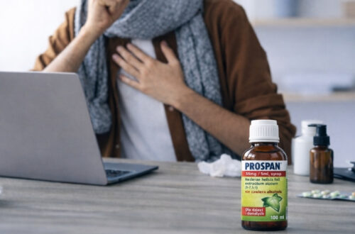 Osoba sa kašljem sedi za laptopom dok je na stolu Prospan sirup za produktivni kašalj