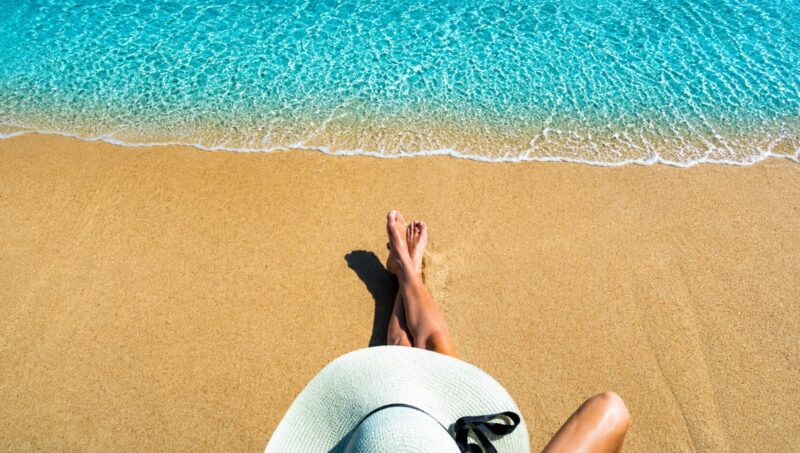 Pogled iz prvog lica na osobu koja sedi na peščanoj plaži pored mora tokom letnjeg dana