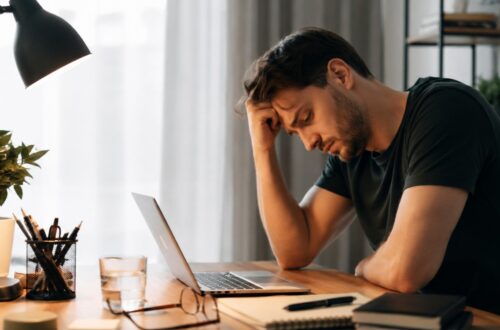 Muškarac sedi za radnim stolom ispred laptopa, drži se za čelo i izgleda mentalno iscrpljeno dok radi u večernjim satima.