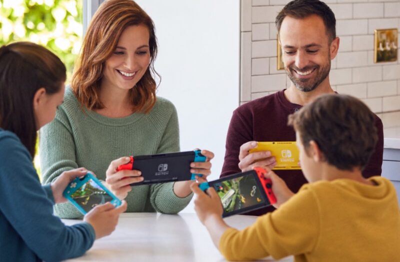 Porodica sedi za stolom i zajedno igra Nintendo Switch konzole u svetlom kućnom enterijeru